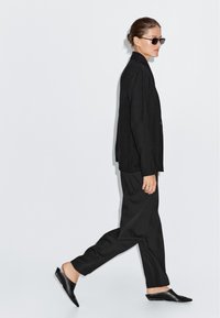 Vrouw die in een zwarte pak loopt met spitse leren instappers en kleine zwarte zonnebril tegen een effen lichte achtergrond.