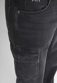 Pantalones cargo de mezclilla negra con un bolsillo de parche texturizado que presenta un sutil diseño de patrones y costuras en contraste. Incluye herrajes metálicos.