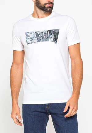 Uomo che indossa una maglietta bianca con stampa collage in bianco e nero di una partita di basket, abbinata a jeans blu, in piedi su sfondo bianco.