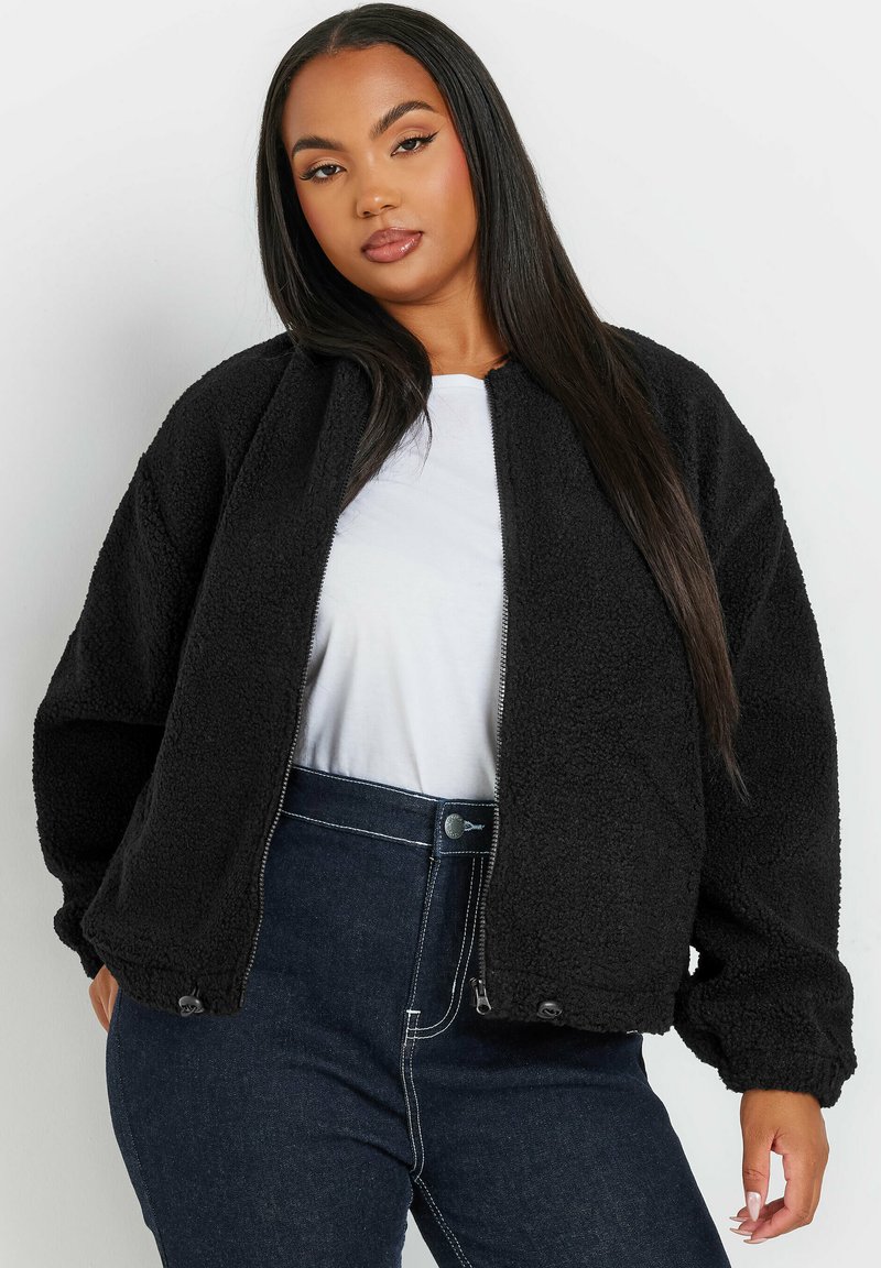 Czarna polarowa kurtka z zamkiem z przodu, ściągaczami przy rękawach i prostokątnym krojem. Nosi ją włożoną na białą koszulę, w zestawieniu z ciemnoniebieskimi dżinsami.