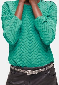 Green long-sleeve top with a zigzag pattern, featuring a round neckline with three black buttons, paired with black leather pants and a silver belt.