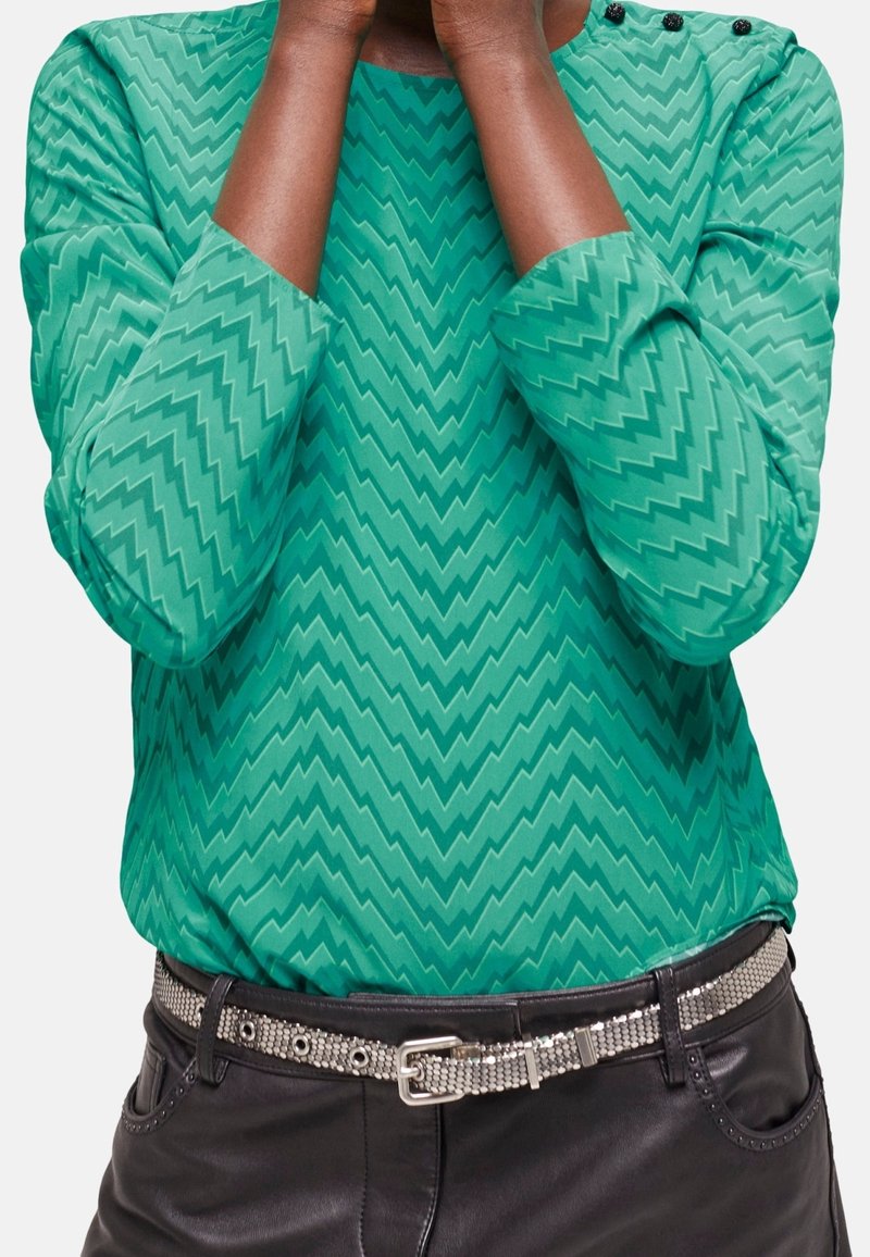Zielona bluzka z długim rękawem w zygzakowaty wzór, z okrągłym dekoltem i trzema czarnymi guzikami, zestawiona z czarnymi skórzanymi spodniami i srebrnym paskiem.