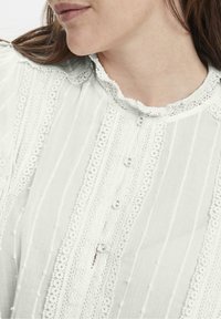 Blusa bianca con colletto arricciato, tessuto a trama con righe verticali, dettagli in pizzo e chiusura con bottoni.