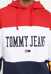 Felpa con cappuccio caratterizzata da blocchi di colore rosso, bianco e blu navy. Testo frontale con la scritta "TOMMY JEANS" in blu navy, con dettagli in accent rosso sotto.
