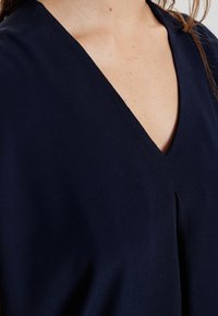 Granatowa bluzka z dekoltem w serek, plisowanym detalem z przodu oraz miękką, gładką fakturą. Brak widocznych wzorów czy ozdób.