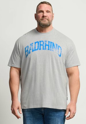 Bärtiger Mann mit kurzen Haaren, der ein hellgraues "BADRHINO" T-Shirt und blaue Jeans trägt, steht vor einem schlichten hellen Hintergrund.
