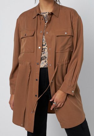 Personne portant une longue veste-chemise marron à boutons avec poches à rabat par-dessus un haut à motifs et un pantalon noir, avec des bagues à la main gauche.