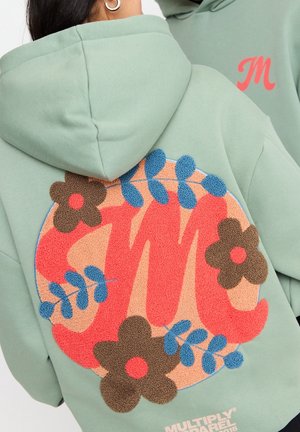 Person iført salviegrøn hættetrøje med store, teksturerede koralrøde "MJ"-bogstaver og brune blomster med blå blade på bagsiden.