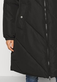 Svart vadderad rock med diagonala sömmar, en silverfärgad dragkedja, smyckade fickor och manschetter. Normal passform och texturerat material.