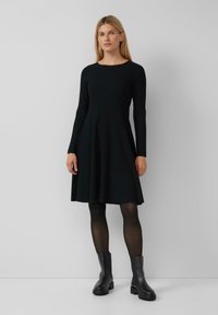 Robe noire à manches longues côtelées avec une jupe évasée, un col rond et une texture lisse, associée à des bottines noires.