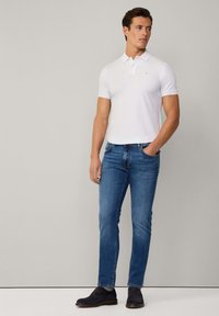 Camisa polo blanca de manga corta con cuello, combinada con jeans de mezclilla azules. La camisa tiene un pequeño logo en el pecho. Los zapatos son oscuros.