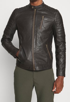 Mand iført en tætsiddende sort læderjakke med lynlåslommer foran og på brystet, stående med den ene hånd i lommen mod en ensfarvet baggrund.