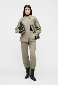 Γυναίκα που φοράει μπεζ υπερμεγέθη σακάκι και παντελόνι σετ, μαύρο ζιβάγκο, κρατώντας μαύρη τσάντα και φοράει μαύρα μποτάκια.