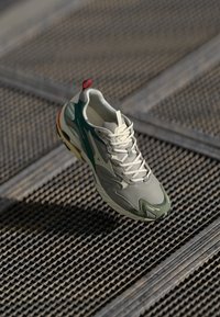 Pantofi sport în bej și verde, cu un superior din plasă, talpă texturată și tab de călcâi roșu, concepuți pentru confort și suport.