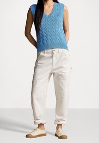 Žena v modré bezrukávové svetrové vestě s pleteným vzorem, off-white kalhotách s rolled-up a skvrnami od barvy, a béžových šn ěrovacích teniskách.