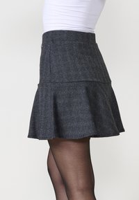 Jupe en chevrons gris avec une taille ajustée et un bas évasé, présentant un ourlet froncé et une texture lisse. Des boutons ajoutent un détail visuel.
