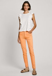 Bijela majica s volanima, u kombinaciji sa suženim narančastim hlačama. Model nosi svijetlosmeđe sandale. Jednostavan, ležeran outfit.