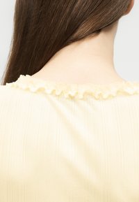 De longs cheveux bruns tombent sur un cou pâle portant un haut jaune clair avec un détail de col à volants doux.