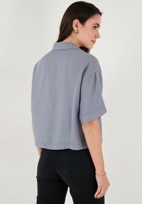 Cropped gray collared shirt made from textured fabric with rolled sleeves, featuring a relaxed fit and a straight hem at the back.