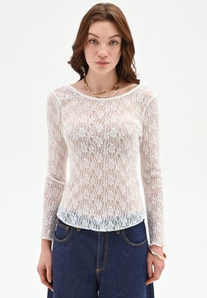 Giovane donna che indossa una maglia bianca a maniche lunghe in pizzo floreale e jeans blu a vita alta, con accessori come collana e braccialetto dorati.