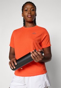 Oransje kortermet T-skjorte med en blå logo, sammen med hvite shorts. Holder en svart sylinderformet vannflaske. Enkelt og rent design.