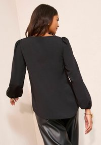 Bluza neagră cu mâneci bufante, decolteu rotund și textură netedă. Asortată cu pantaloni negri lucioși, evidențiind o croială lejeră.