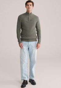 Man in olijfgroene geribde ritstrui, lichtblauwe jeans en zwarte leren schoenen, staande tegen een effen achtergrond.