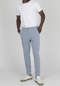 Svetle modré kalhoty na míru s hladkým povrchem, bočními kapsami a zapínáním na knoflík, doplněné bílým tričkem s krátkým rukávem a teniskami.