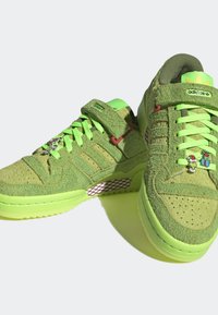 Zelené texturované tenisky Adidas s neonově zelenými tkaničkami, vánočními ozdobnými uzlíky a suchým zipem na jednoduchém světlém pozadí.