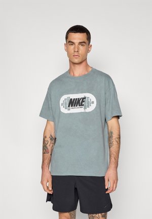 T-shirt en coton gris avec un grand logo Nike noir et blanc centré. Manches courtes et coupe décontractée. Porté avec un short de sport noir.