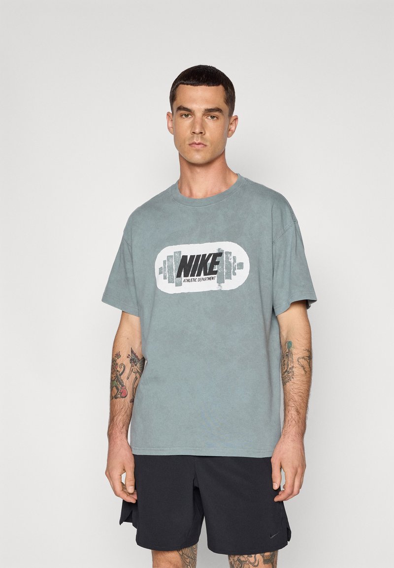 Šedé bavlněné tričko s velkým černobílým logem Nike uprostřed. Krátké rukávy a volný střih. Nosí se s černými sportovními šortkami.