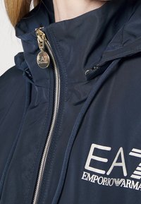 Marinblå vattentät jacka med hög krage, guldfärgad dragkedja och EA7 Emporio Armani-logotyp. Har justerbar huva och slät textur.