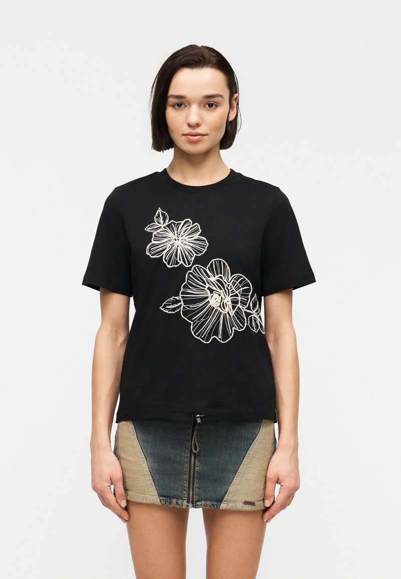 Femme portant un t-shirt noir avec une broderie florale blanche et une mini-jupe en denim bicolore avec une fermeture éclair à l'avant.