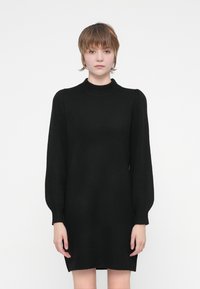 Melna adīta kleita ar augstu apkakli, garām puķu piedurknēm un brīvu piegriezumu, ar ribotām aprocēm un apakšmalu, lai pievienotu tekstūru.