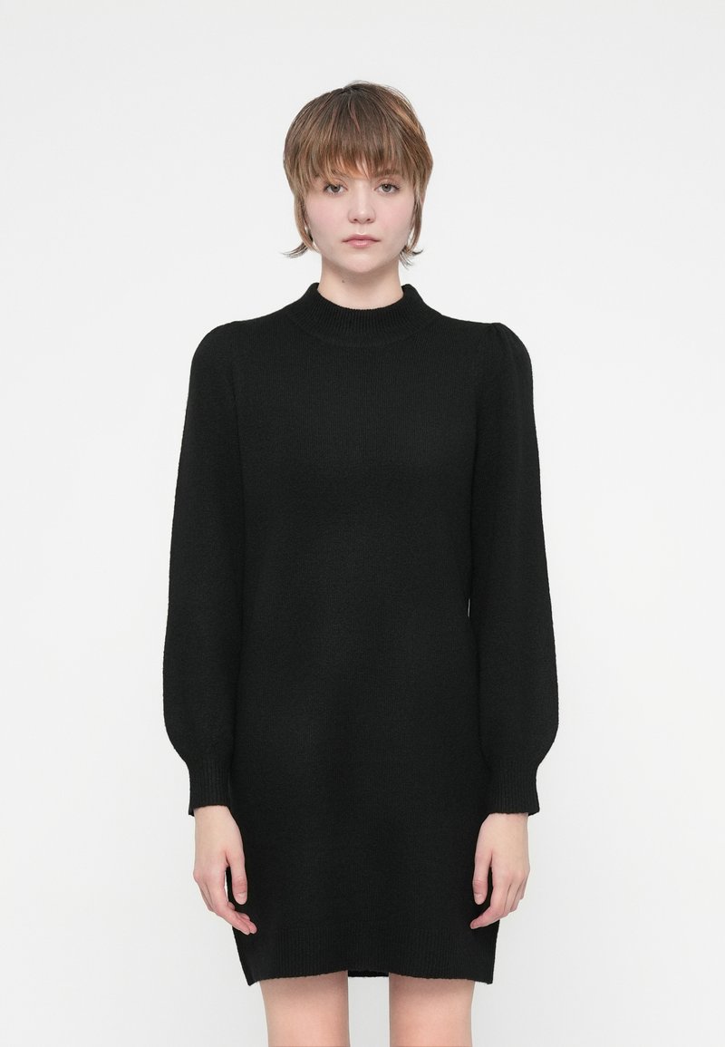 Melna adīta kleita ar augstu apkakli, garām puķu piedurknēm un brīvu piegriezumu, ar ribotām aprocēm un apakšmalu, lai pievienotu tekstūru.