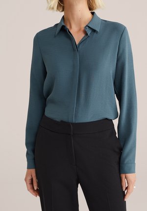 Vrouw draagt een teal-kleurige blouse met lange mouwen en kraag, ingestopt in hoge zwarte broek, staand tegen een effen achtergrond.