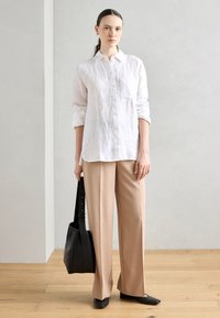 Camicia button-up bianca in lino sovrapposta a pantaloni beige a gamba larga, abbinata a scarpe flat nere e una borsa a spalla nera. Design minimalista.