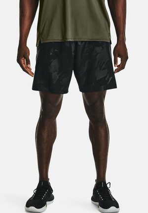 Homme portant un short de sport noir, un t-shirt vert olive et des chaussures de course noires sur un fond blanc uni.