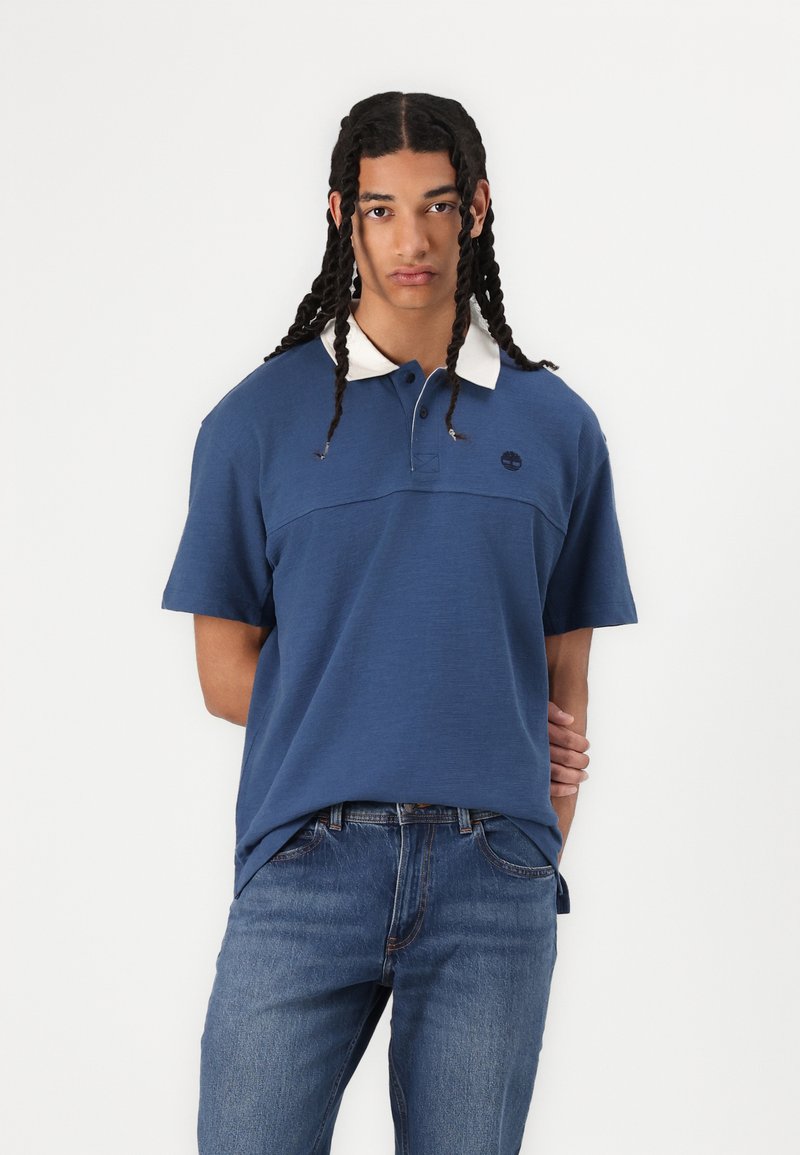 Polo shirt bleu marine avec un col blanc, à manches courtes, avec une patte de boutonnage à l'avant et un logo sur la poitrine. Porté avec un jeans bleu.