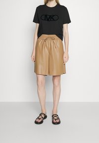 Camiseta de algodón negra con un logotipo de terciopelo, pantalones cortos de cuero sintético color marrón con una cinturilla elástica y sandalias negras de tiras en un entorno minimalista.