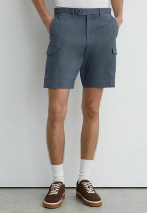 Mand iført blå cargoshorts, hvide crew-sokker og brune sneakers, stående med hænderne i lommerne op ad en helt hvid væg.