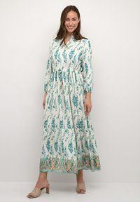 Lång, flödande klänning i krämfärg med tealblommönster, samlad midja och en lager-skirt. Har lång ärmar och en V-ringning.