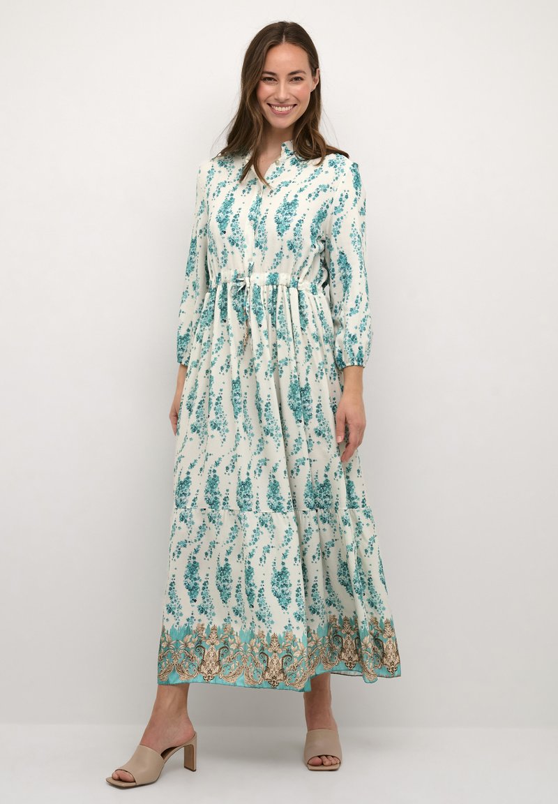 Lång, flödande klänning i krämfärg med tealblommönster, samlad midja och en lager-skirt. Har lång ärmar och en V-ringning.