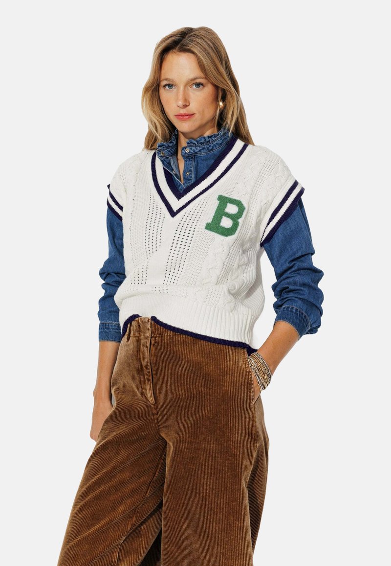 Gilet en tricot blanc à col en V, accent "B" vert, porté par-dessus une chemise en denim à col volé et un pantalon large en velours côtelé marron.