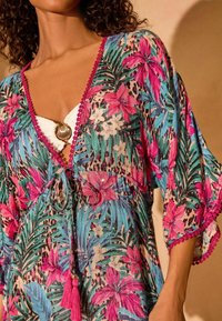 Kimono con stampa floreale in un vivace rosa, verde e blu, caratterizzato da una chiusura a fiocco, dettagli con nappine e un bordo scallop lungo il colletto.