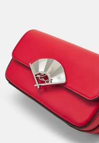 Lille rød læderhåndtaske med en sølvfarvet fanformet lås, der har et krydset linjemønster og kursiv detaljer på klappen.