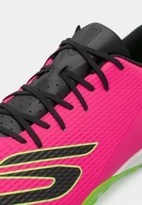 Κοντινό πλάνο από αθλητικό παπούτσι σε νέον ροζ χρώμα με μαύρες αλυσίδες, μαύρη γλώσσα και μαύρο λογότυπο που περιβάλλεται από κίτρινο-πράσινο σε υφασμάτινη επιφάνεια.