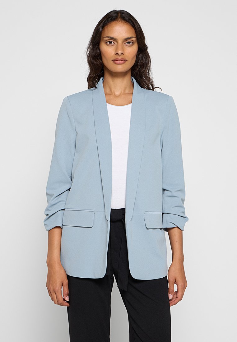 Blazer bleu clair en tissu lisse avec un col châle, des manches retroussées et deux poches avant. Porté sur un top blanc et un pantalon noir.