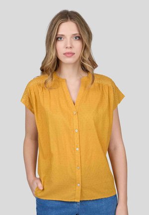 Blouse jaune moutarde à manches courtes avec un devant à boutons, en tissu texturé, et des accents en dentelle sur les épaules, portée avec un jean en denim bleu.