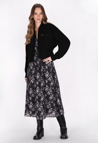 Donna con lunghi capelli mossi, che indossa una giacca nera corta sopra un vestito midi a fiori nero e stivali neri fino al ginocchio, in piedi con una mano sui fianchi.
